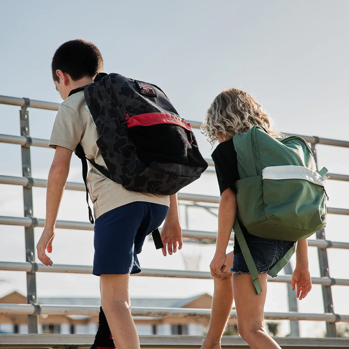 Two kids wearing GORUCK backpacks outdoors, walking up metal bleachers in sunlight