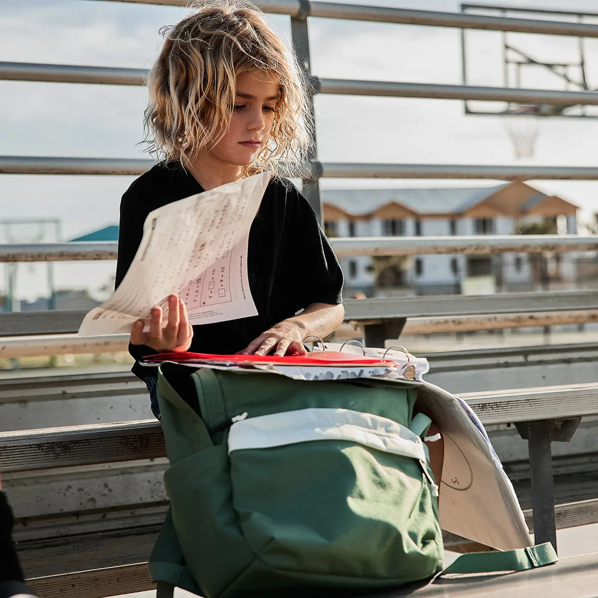 Young child sitting on outdoor bleachers holding papers with open green GORUCK backpack