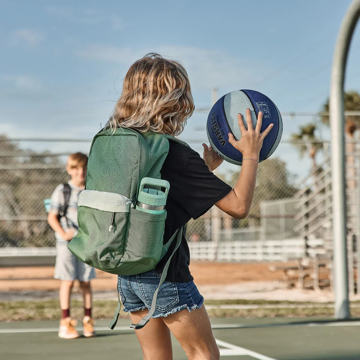 Teen with green GORUCK Kid Ruck backpack playing basketball outdoors on court with blue sky