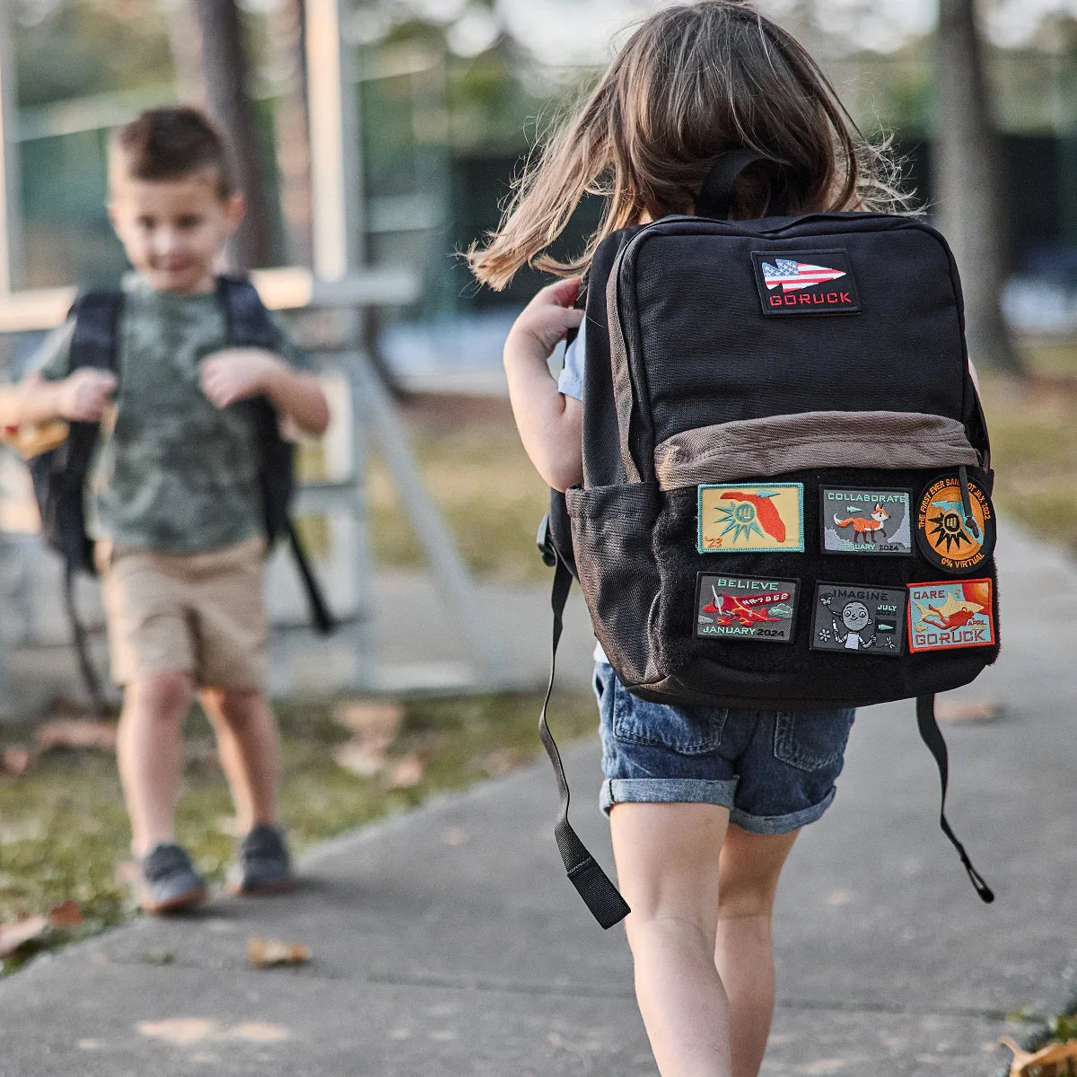 Child wearing black GORUCK Kids Ruck backpack with colorful patches walking outdoors on sidewalk