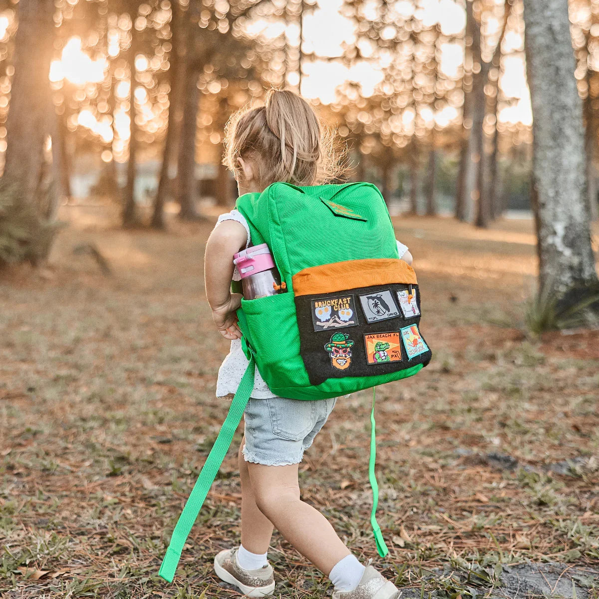 Young child walking outdoors wearing bright green rucksack with colorful patches, carrying a pink water bottle
