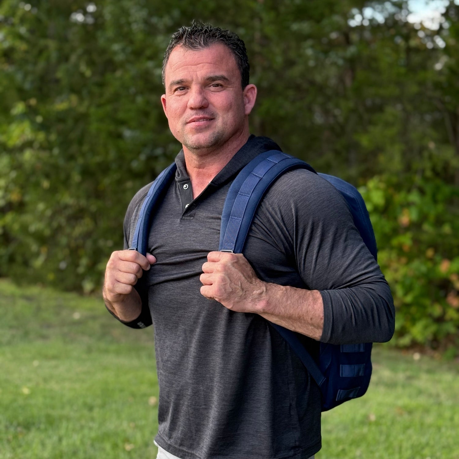 Man standing outdoors with a blue backpack, wearing a dark long-sleeve shirt, and greenery in the background.