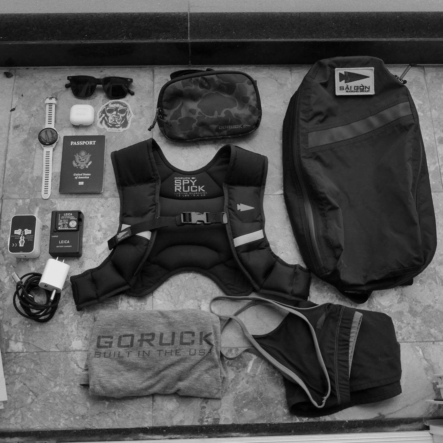 Flat lay of travel essentials: Spy Ruck backpack, Spy Ruck Women's Weighted Vest, clothes, watch, passport, charger, sunglasses, and small accessories.