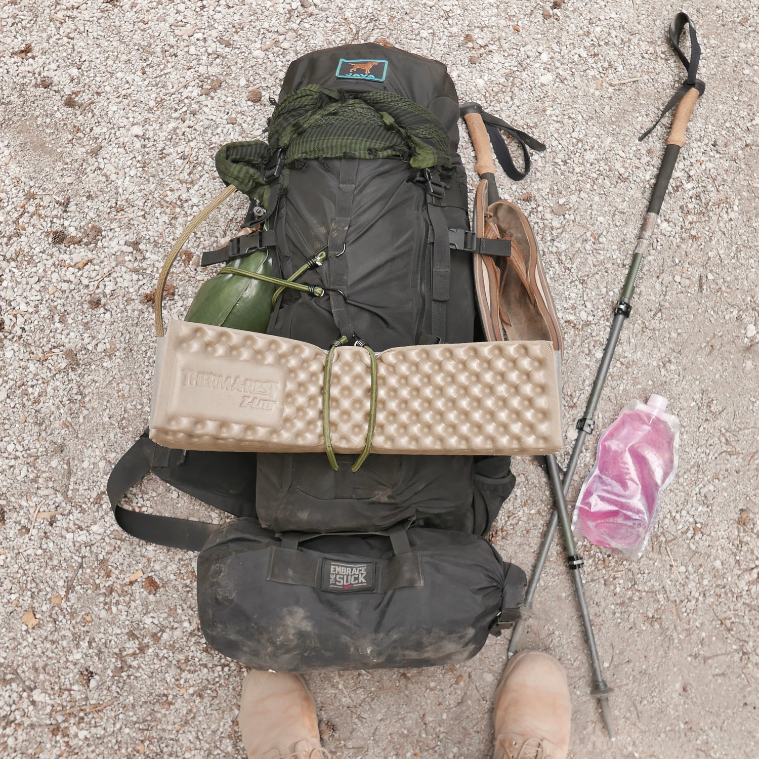 The GR MONSTER Bundle - PREORDER, featuring a packed rucksack, sleeping pad, water bottle, poles, and pink bag on gravel with boots visible.