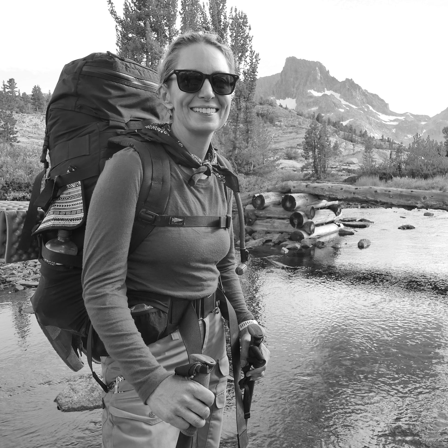 A smiling woman with trekking poles stands by a river, mountains in the background, carrying the GR MONSTER Bundle - PREORDER rucksack.