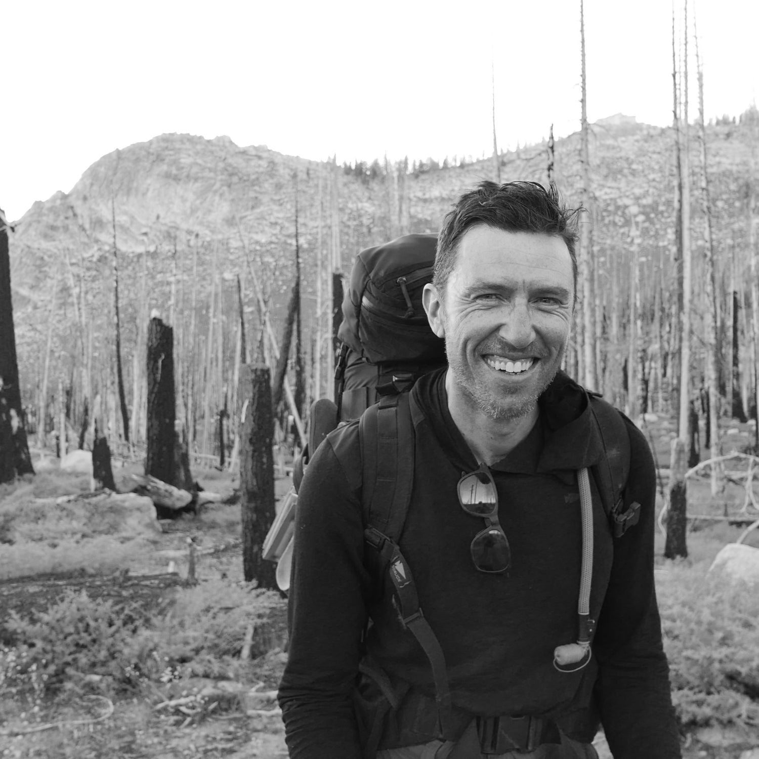 A man carrying the GR MONSTER Bundle - PREORDER smiles while hiking through a forest with charred trees and mountains in the background.