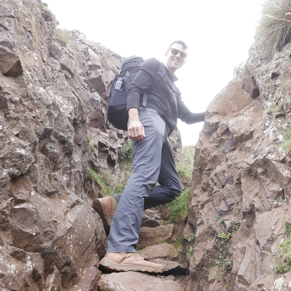 A man with a backpack hikes between rocky cliffs, smiling at the camera on a cloudy day, wearing rugged MACV-2 - Mid Top - Briar + Coyote boots designed for adventure.
