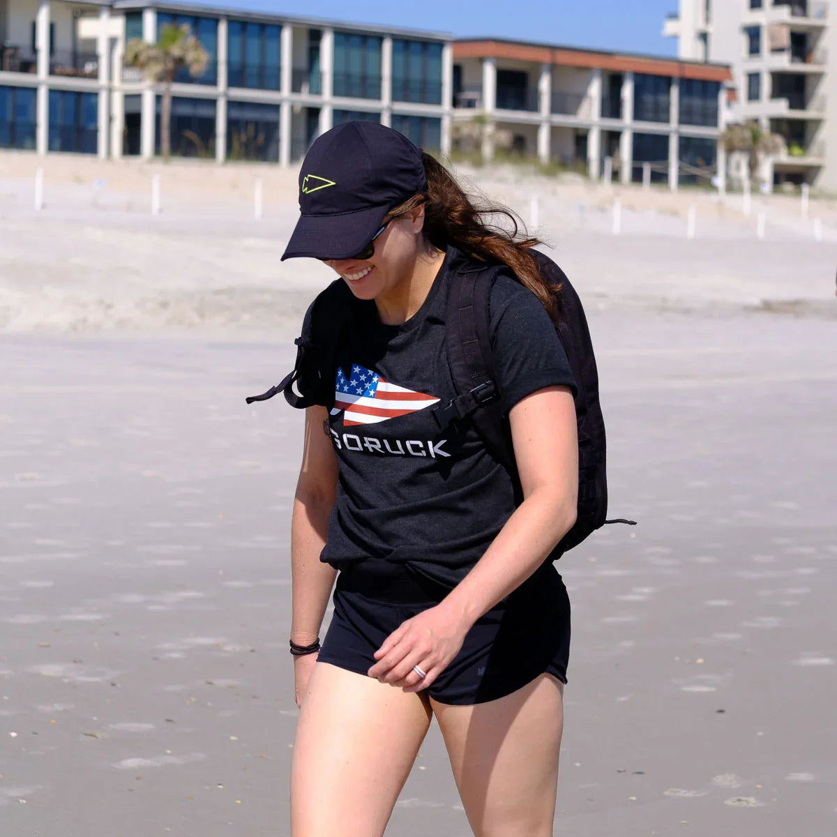 Woman wearing GORUCK black LEAD flag t-shirt and black cap walking on a sunny beach with backpack