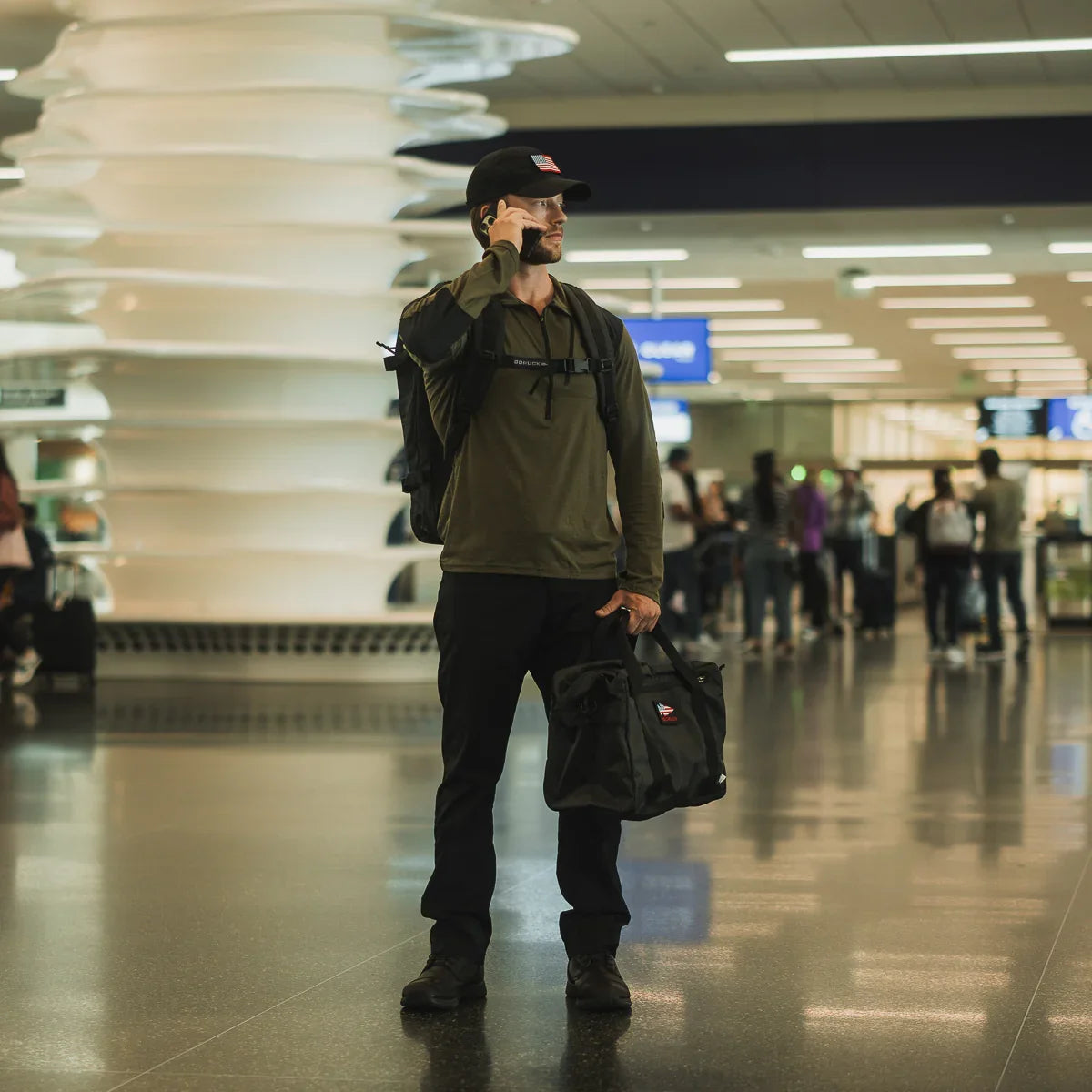 Man in ranger green half-zip merino wool shirt, black pants, and cap, carrying black GORUCK bag and backpack in airport