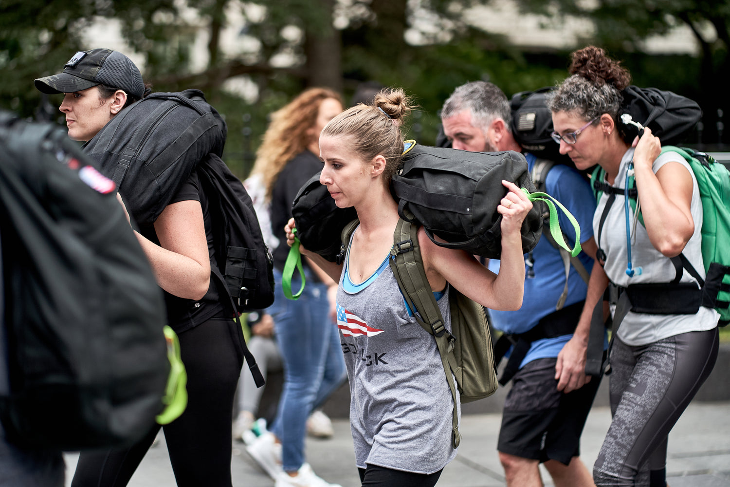 A group of people outdoors carry weighted backpacks on their shoulders during a fitness challenge.