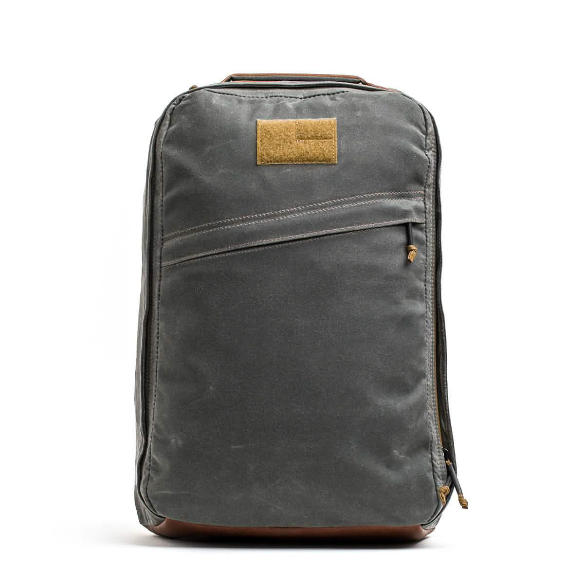 The GR1 USA - Heritage Waxed Canvas is a gray backpack featuring a diagonal zipper pocket and a tan front patch, crafted for timeless style.