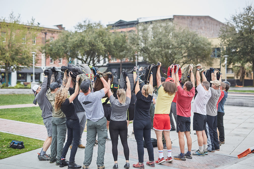 A group of people outdoors raise backpacks over their heads in a circle, facing away from the camera.