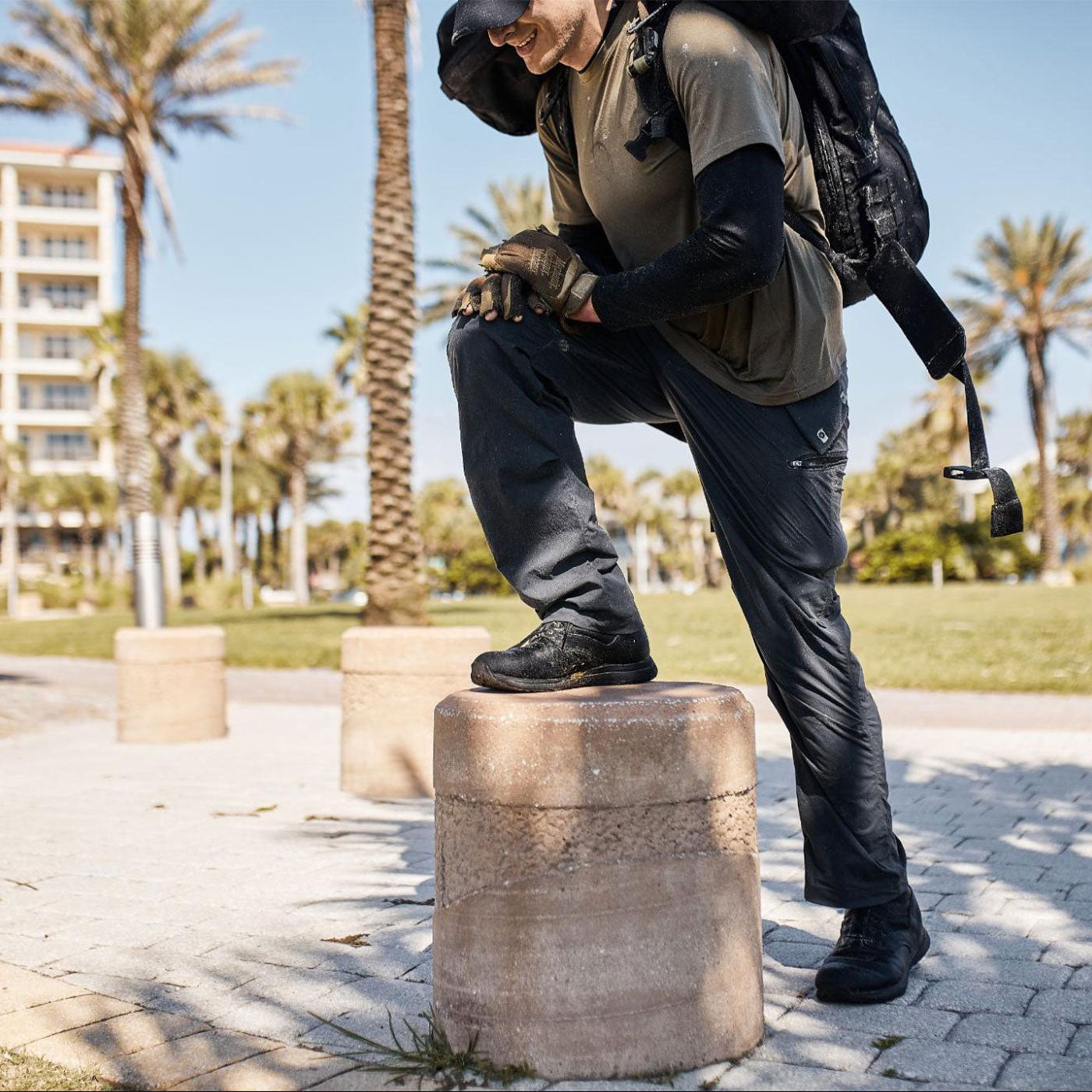 Man in outdoor gear with backpack, one foot on a concrete post, palm trees and buildings in the background.