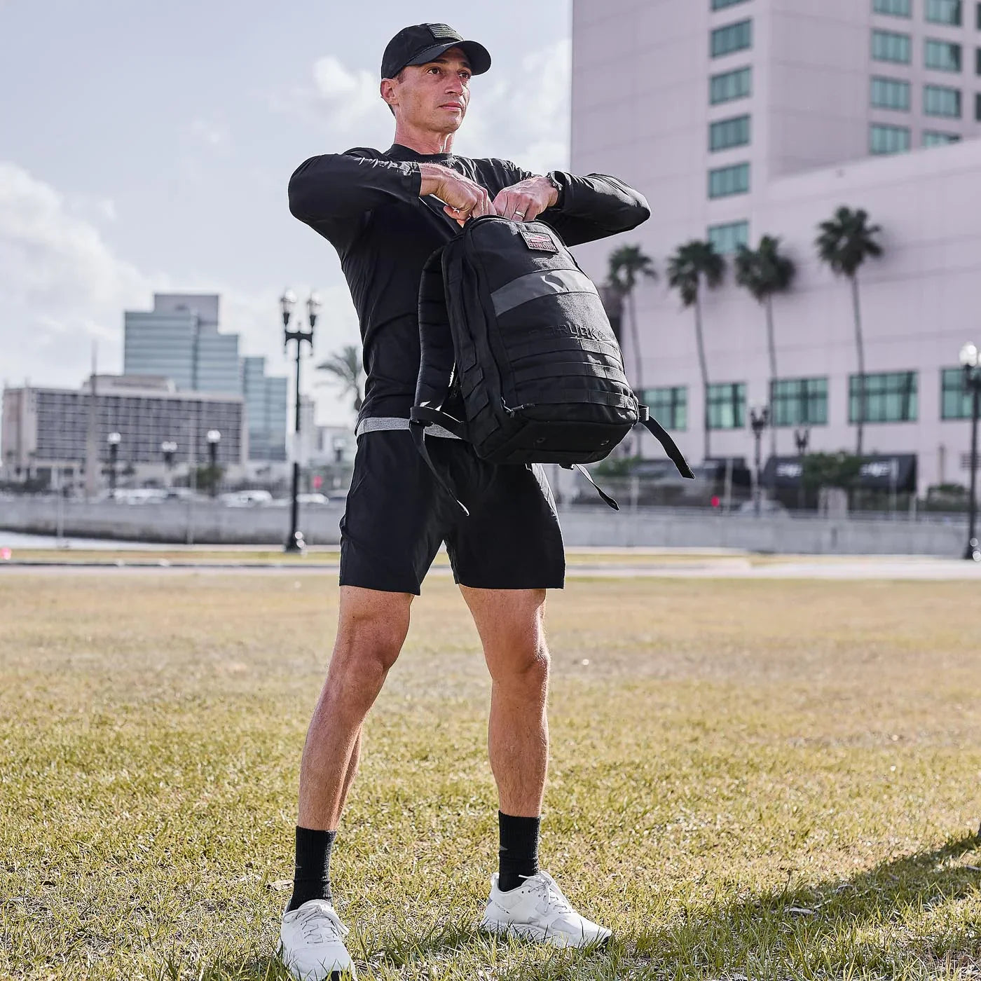 Man wearing black cap, black athletic shorts, and long sleeve performance tee holding black GORUCK backpack in urban park