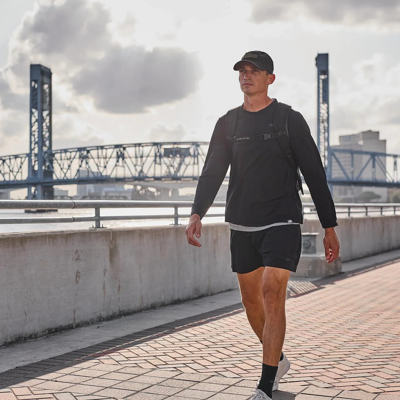 Man walking outdoors wearing black GORUCK long sleeve performance tee, shorts, cap, and backpack near bridge