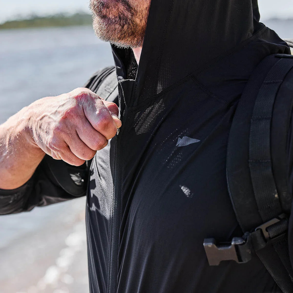 Close-up of a man zipping a black GORUCK performance hoodie with a backpack outdoors near water