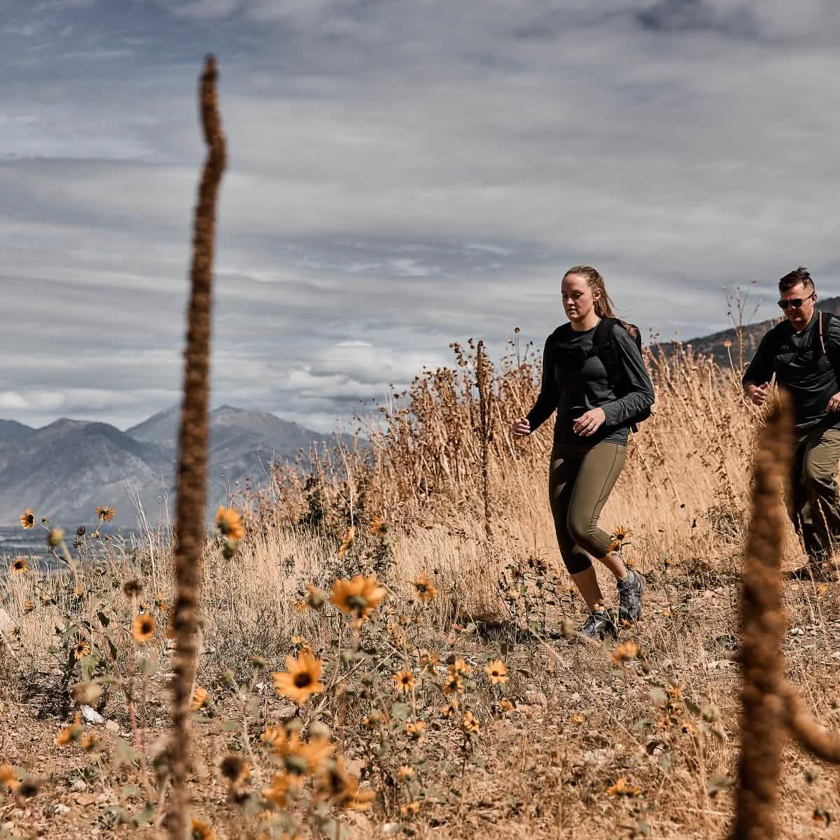 Two hikers walking through a dry, grassy trail with sunflowers and mountains in the background under a cloudy sky