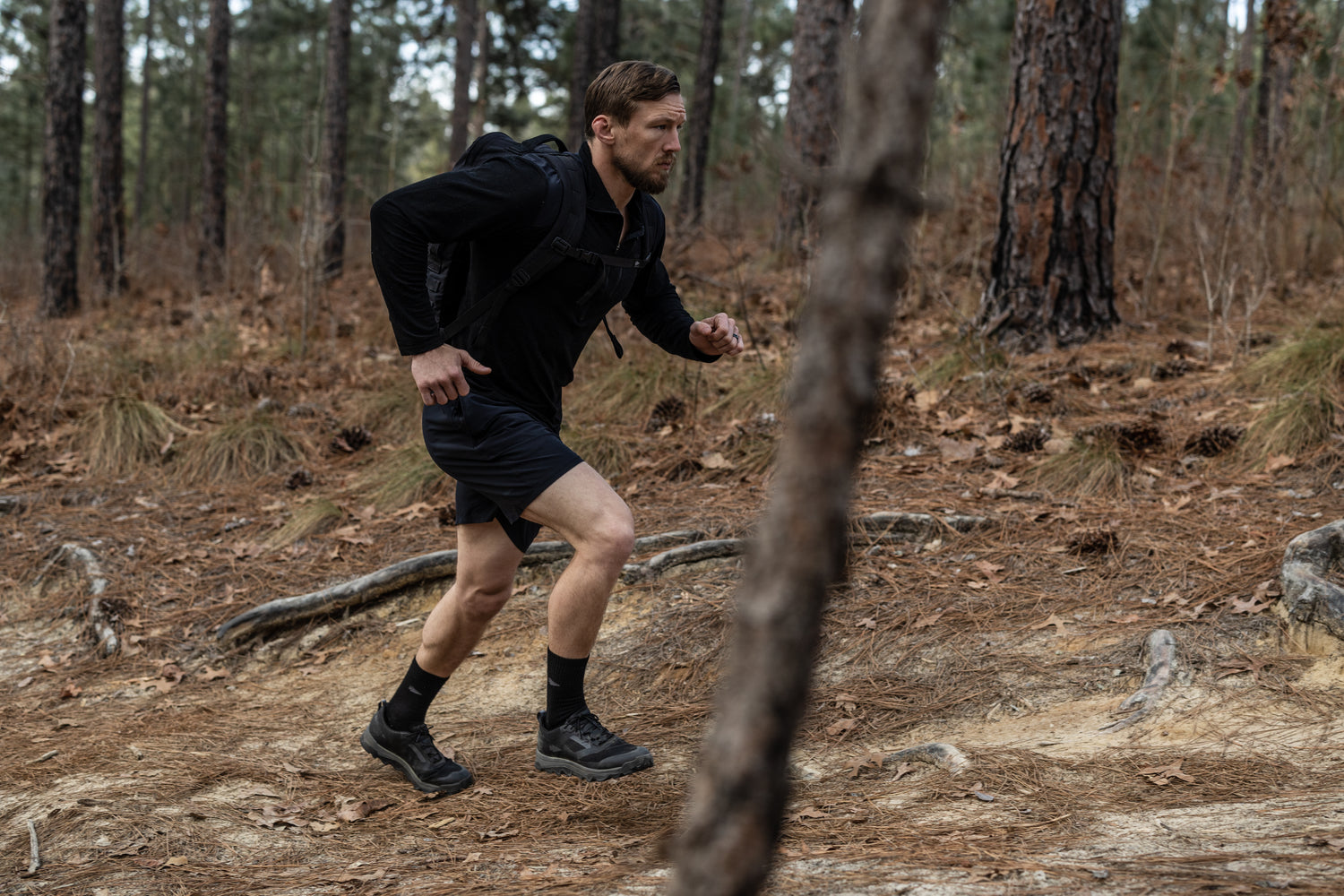 Man wearing a backpack hiking uphill on a forest trail with trees and dry leaves around.