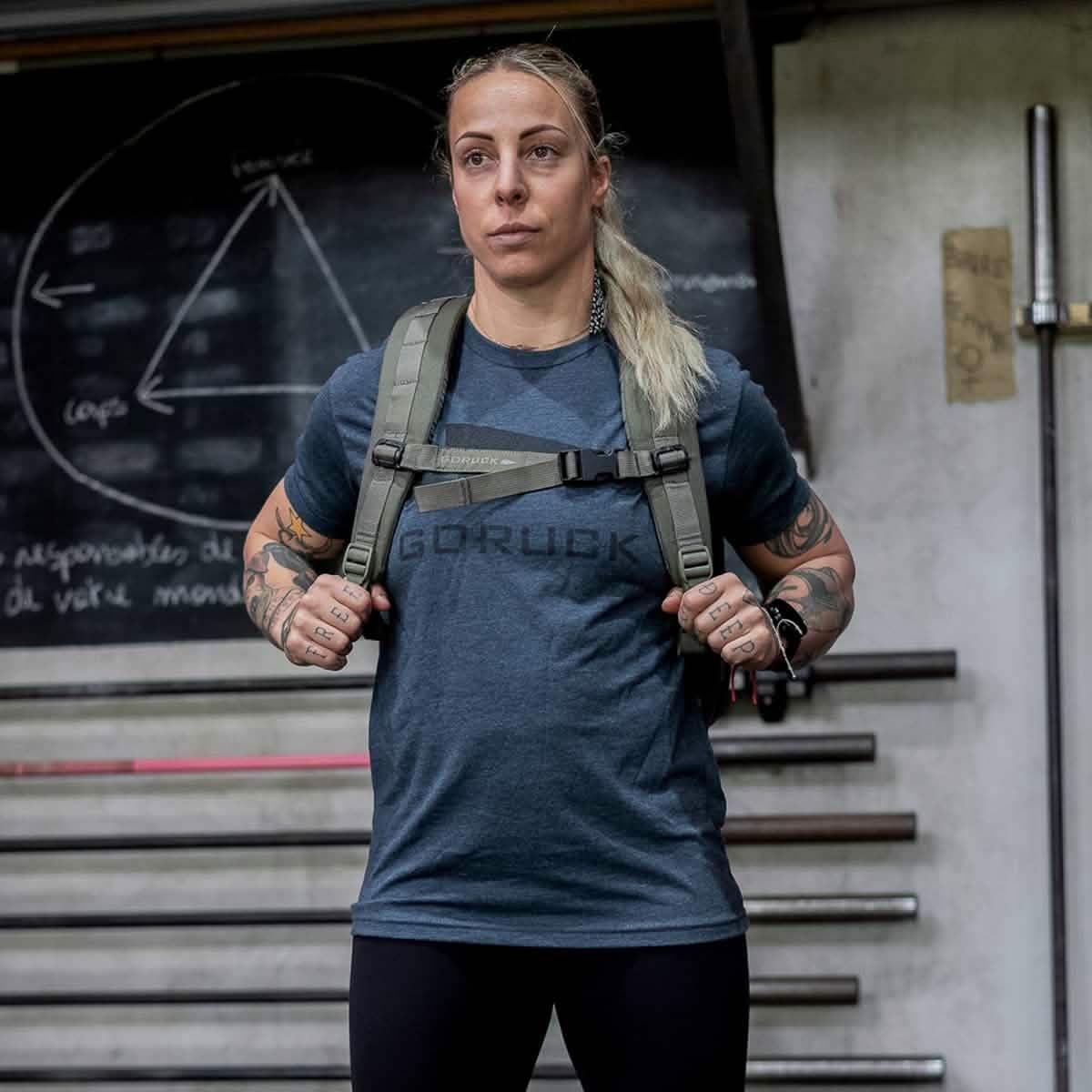 Woman wearing GORUCK navy tee and rucksack in gym with chalkboard and fitness bars