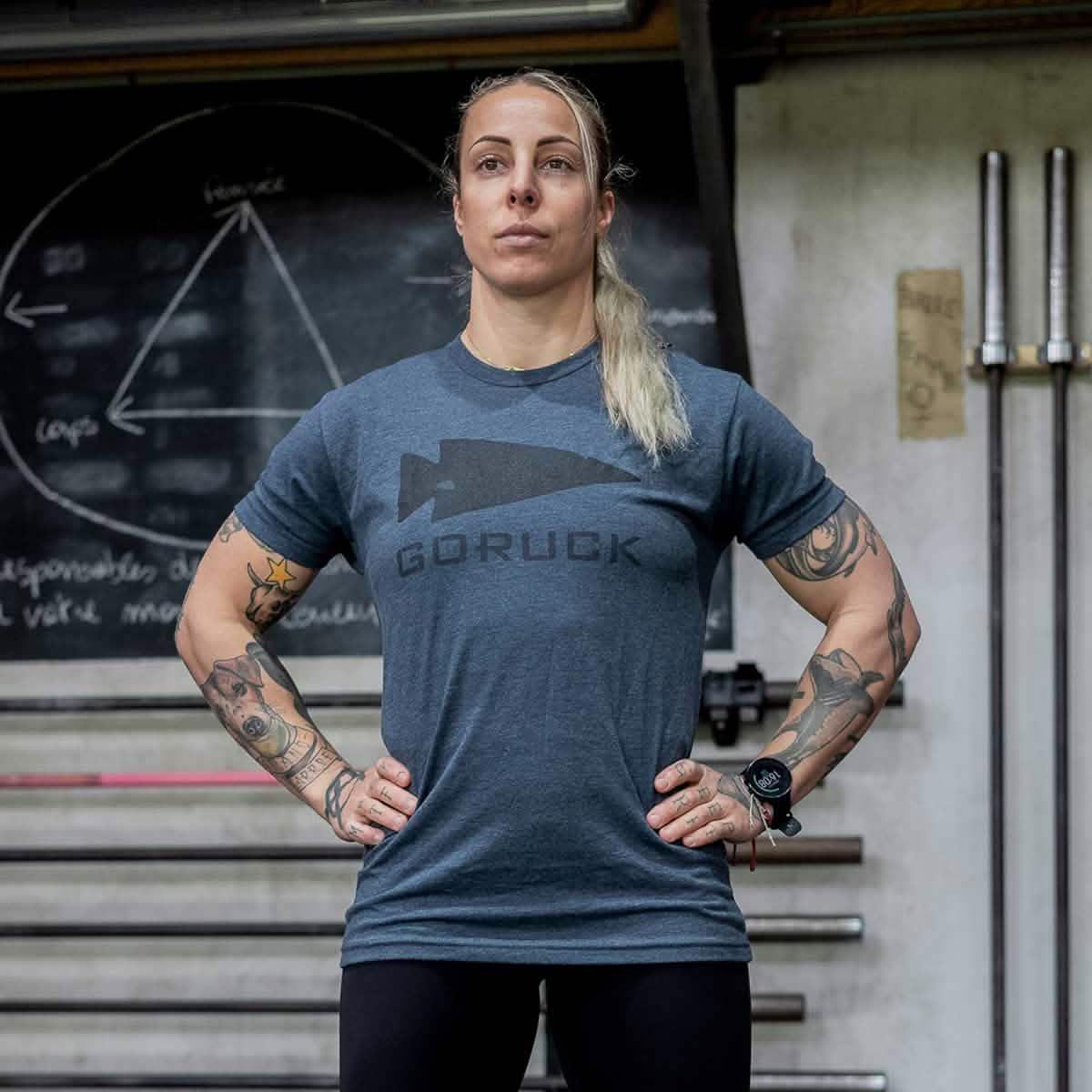 Athletic woman in navy GORUCK Spearhead tee standing in gym, weightlifting bars and chalkboard behind her.