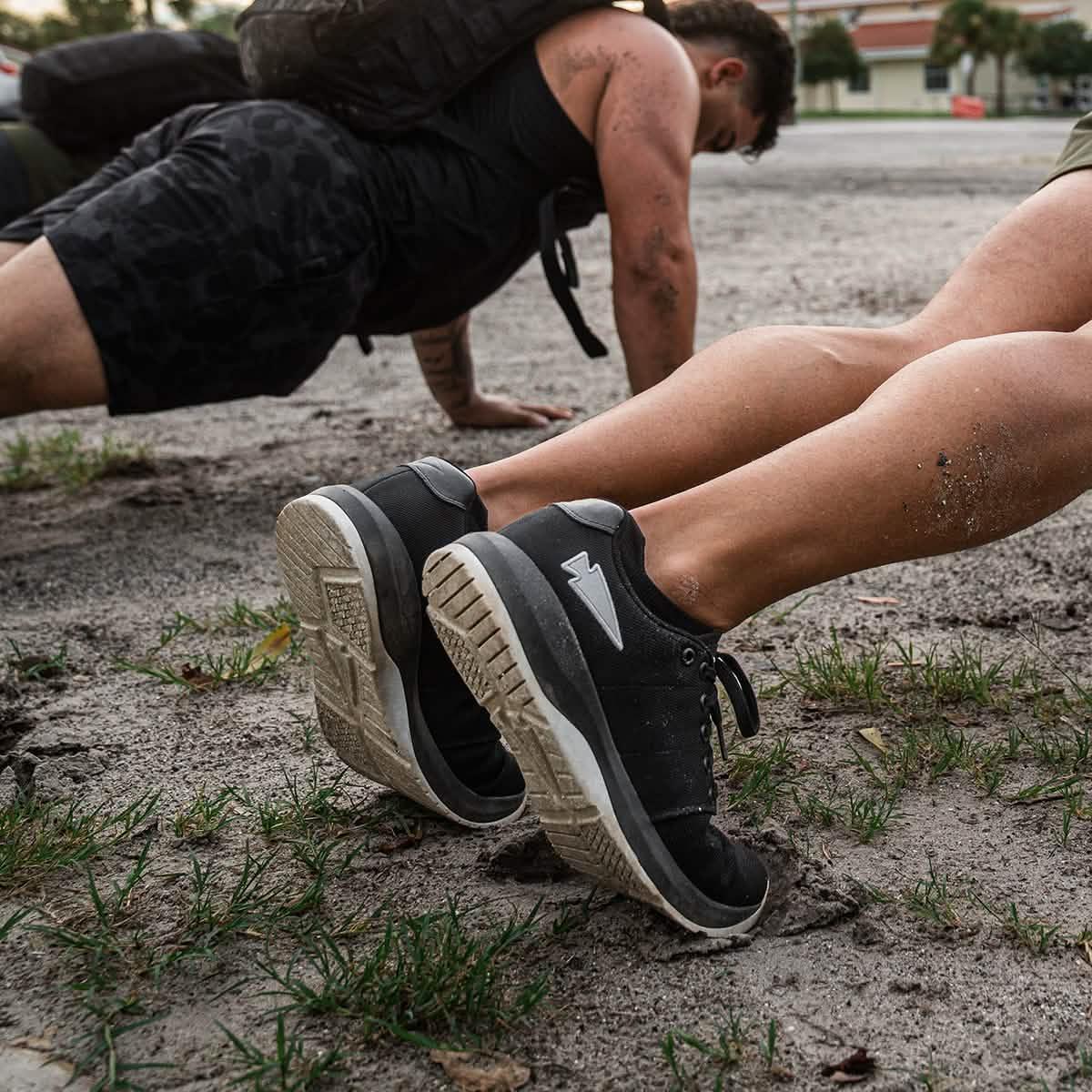 Close-up of black GORUCK ballistic trainers worn during outdoor push-up exercise on dirt ground