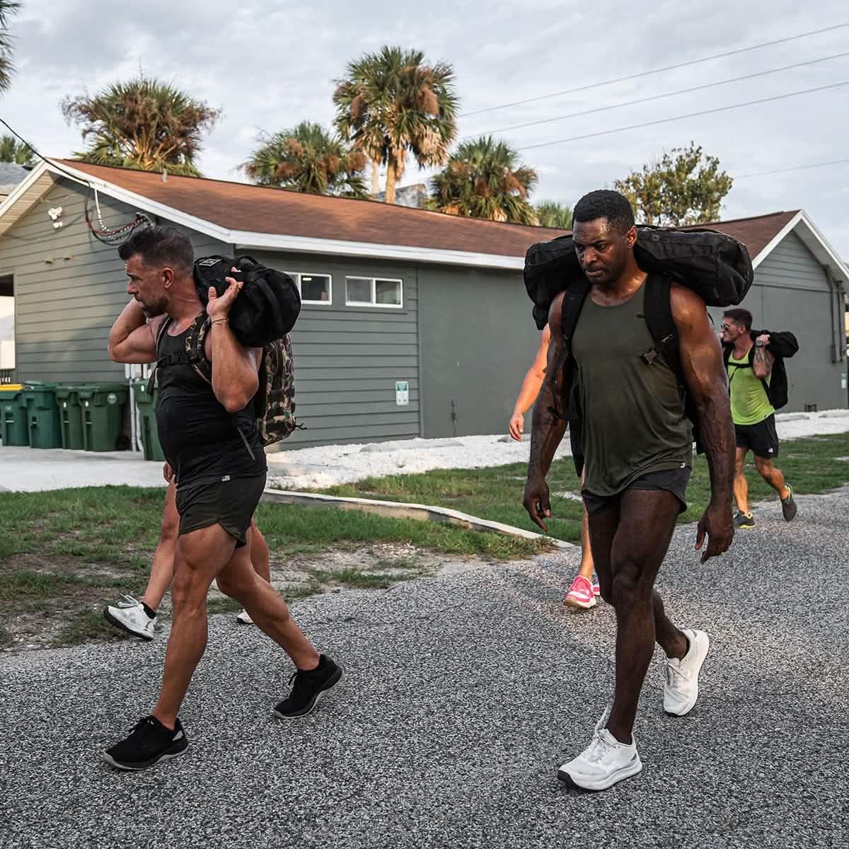 Men carrying weighted rucksacks during outdoor rucking workout near residential buildings