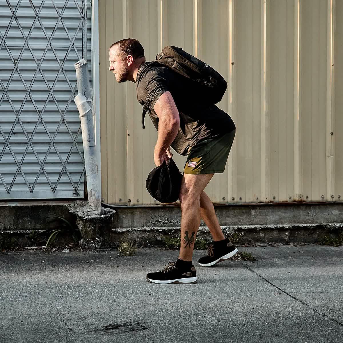 Man wearing GORUCK black ballistic trainers and backpack, outdoors on concrete next to metal siding