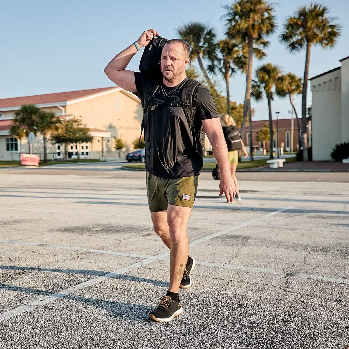 Man rucking outdoors in GORUCK Ballistic Trainers, wearing black shirt, green shorts, backpack.