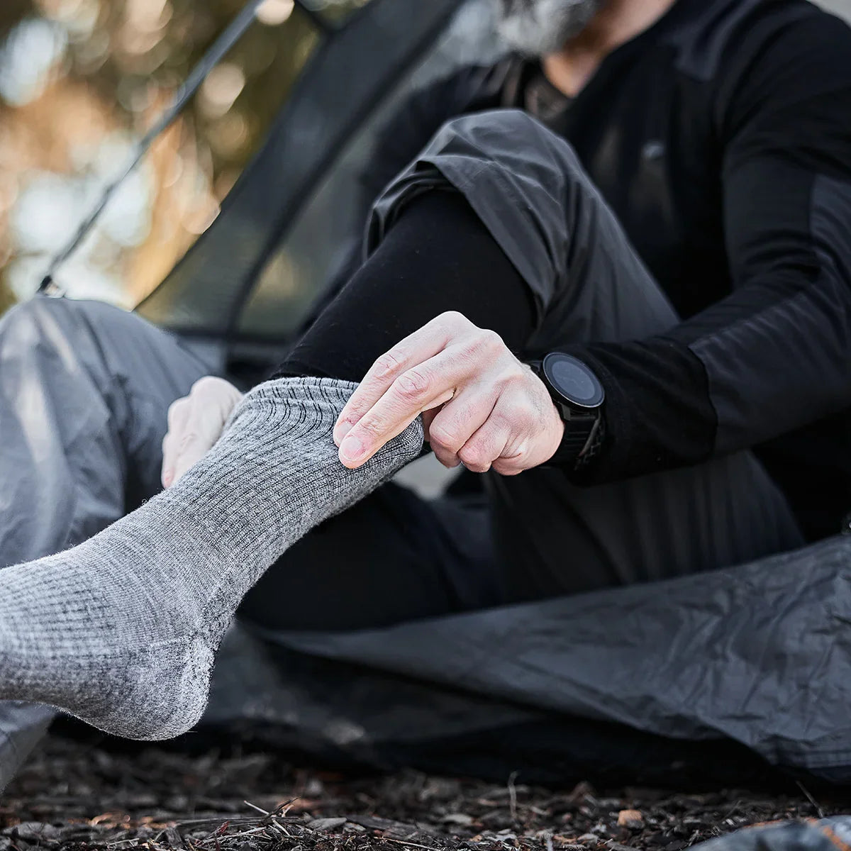 Man outdoors wearing GORUCK merino wool base layer, adjusting gray socks inside tent