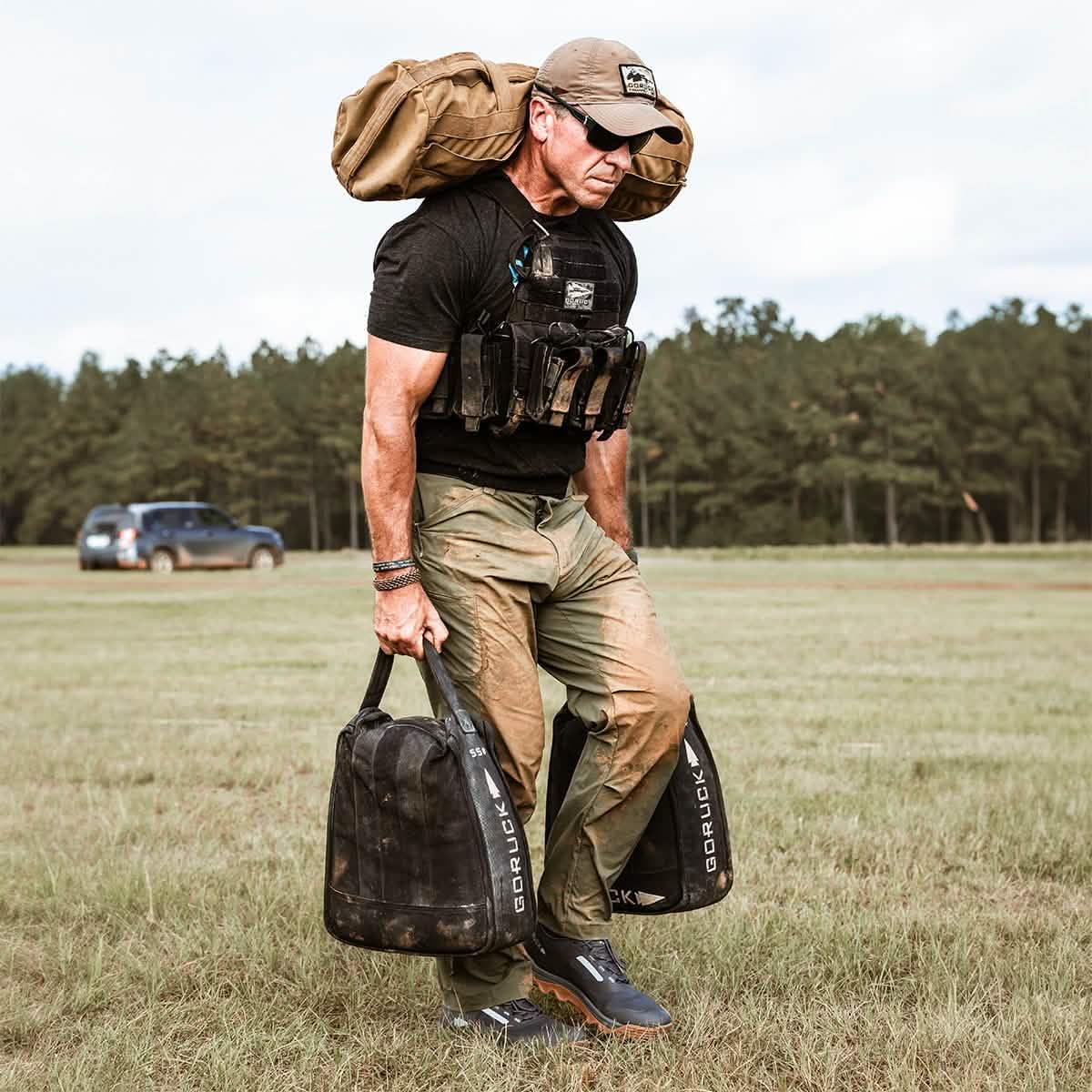 Man in ranger green tactical pants carrying GORUCK weighted bags outdoors in a grassy field with forest background