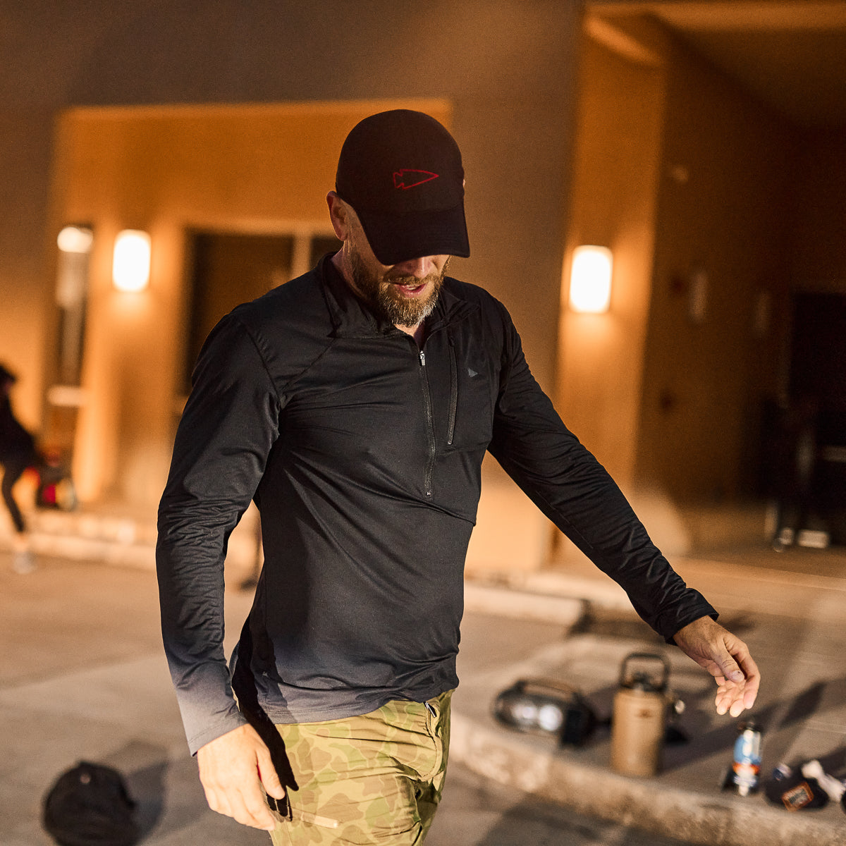 A man wearing the Men's USA Long Sleeve Performance Half Zip - ToughMesh and a cap walks outdoors at night near gym equipment.