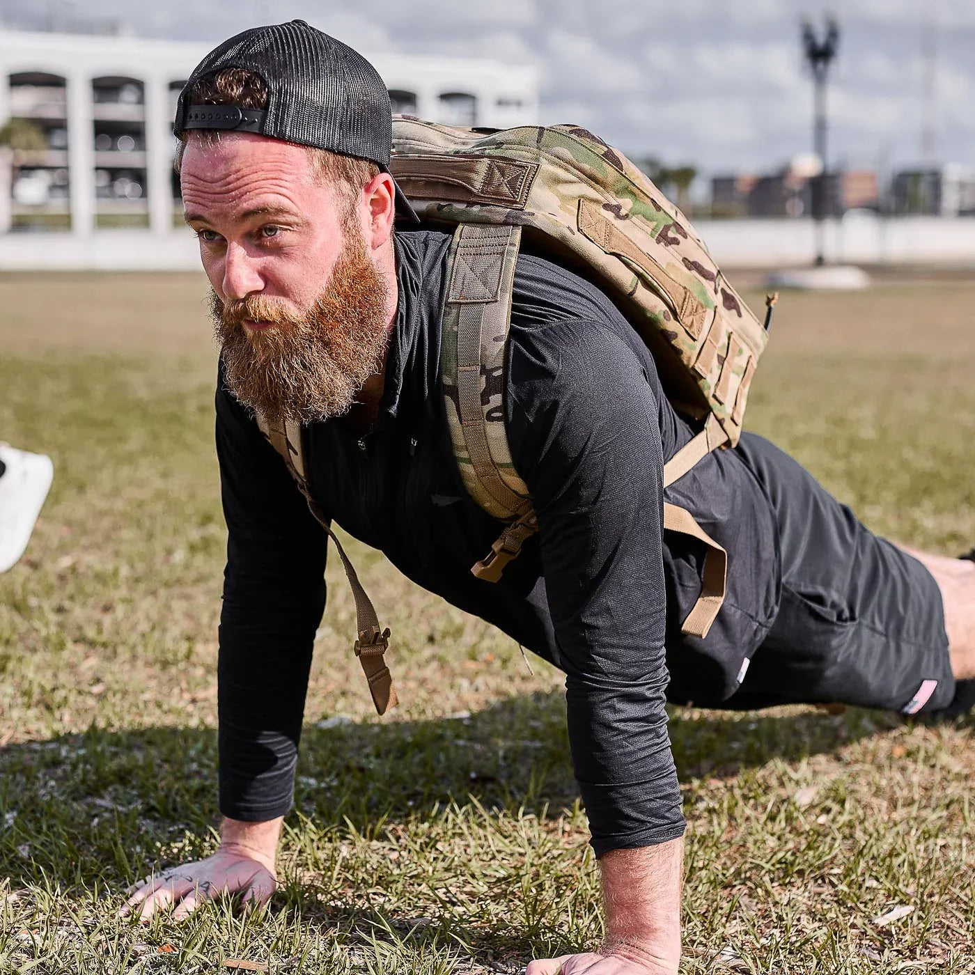 Man doing outdoor push-up in GORUCK long sleeve with camo rucksack, grass field background