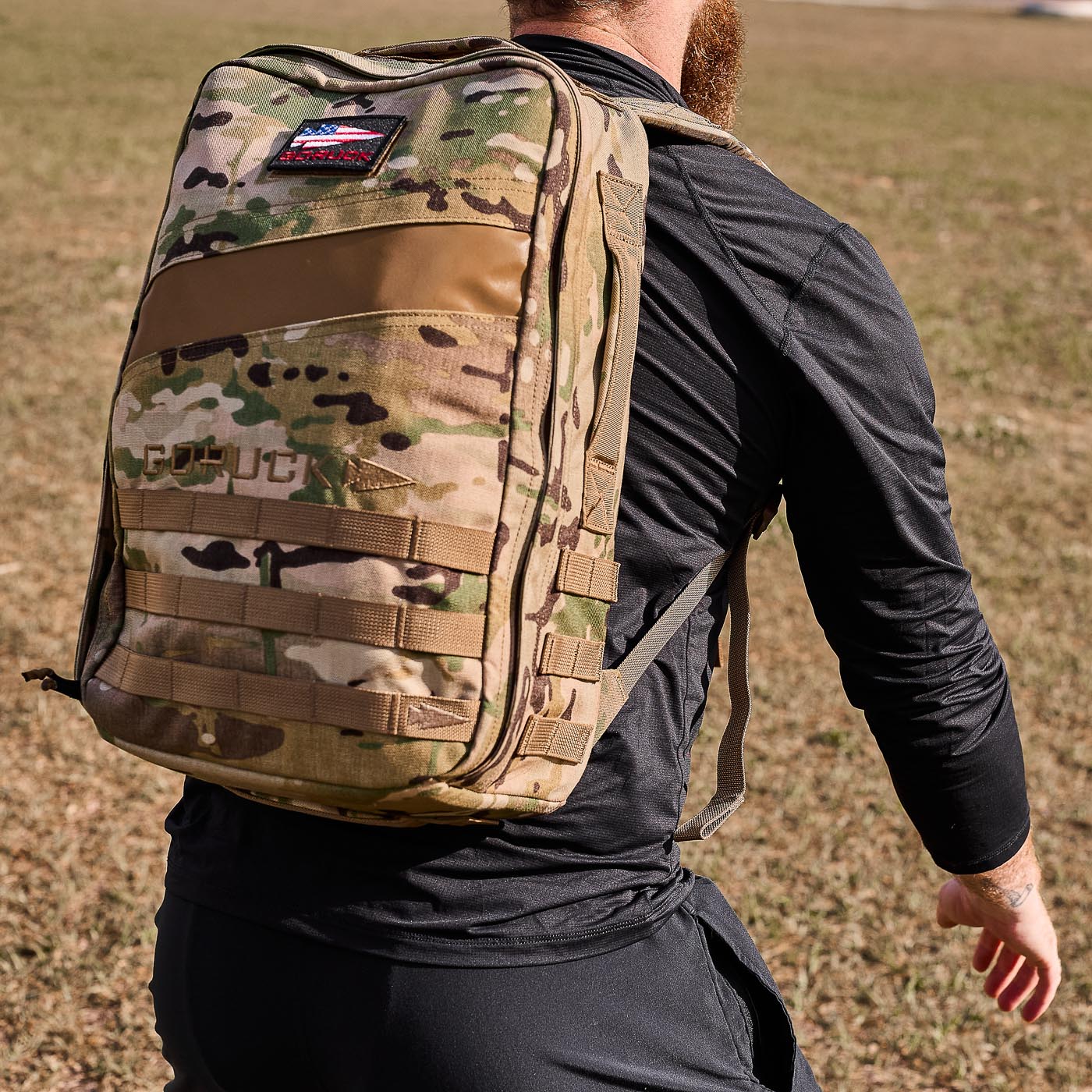 Man wearing the Men's USA Long Sleeve Performance Half Zip - ToughMesh and a camouflage backpack on a grassy field. Black athletic clothing offers featherweight comfort, and the Scars Lifetime Guarantee ensures lasting performance outdoors.