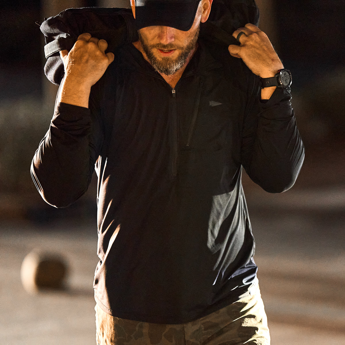 A man in black athletic wear lifts a sandbag outdoors, wearing a cap and camouflage pants. His Men's USA Long Sleeve Performance Half Zip - ToughMesh ensures comfort during intense workouts.
