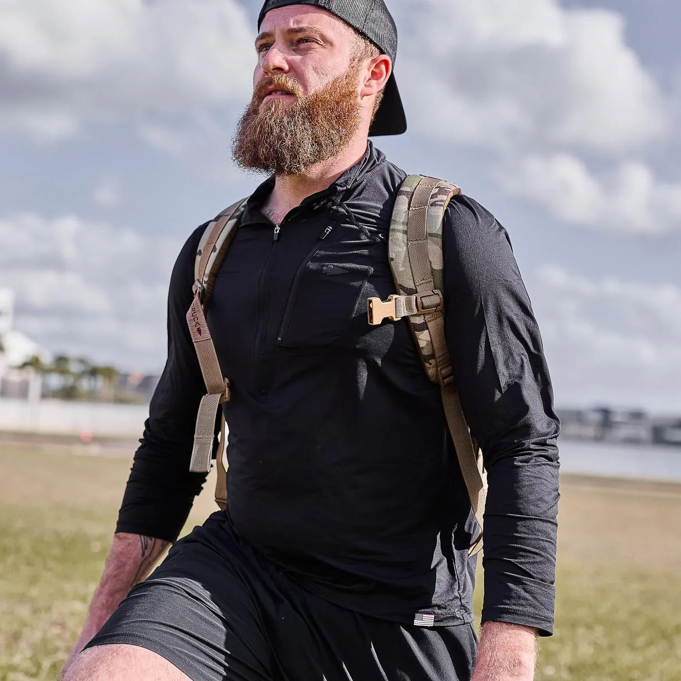 Bearded man wearing black long-sleeve performance half-zip shirt and camo backpack outdoors