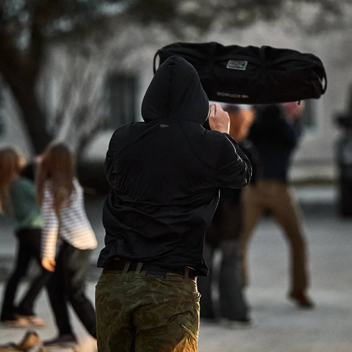 Person in black hoodie and camo pants throwing a black GORUCK weighted bag outdoors with blurred background