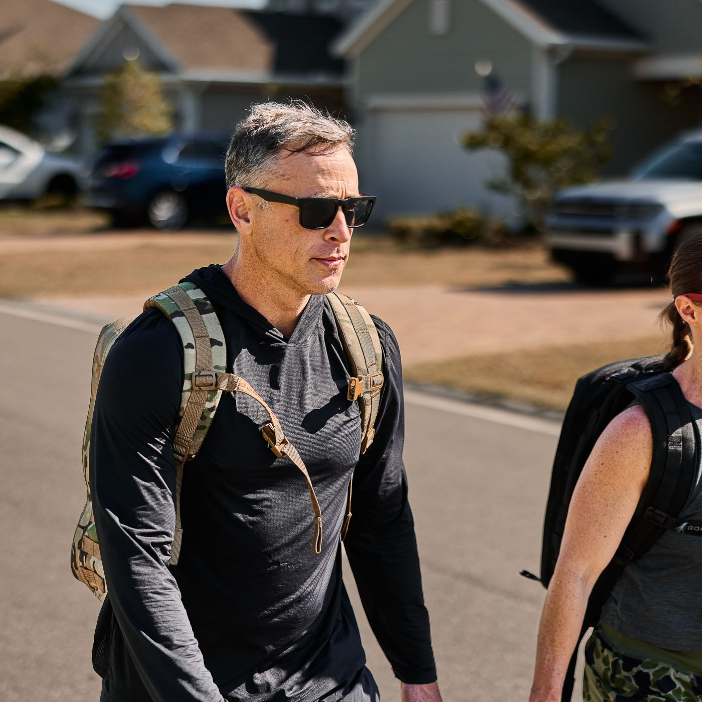 A man wearing the Men's USA Sun Hoodie - ToughMesh with a backpack strolls in a sunny suburban neighborhood, enjoying breathable comfort and UPF 30+ sun protection.