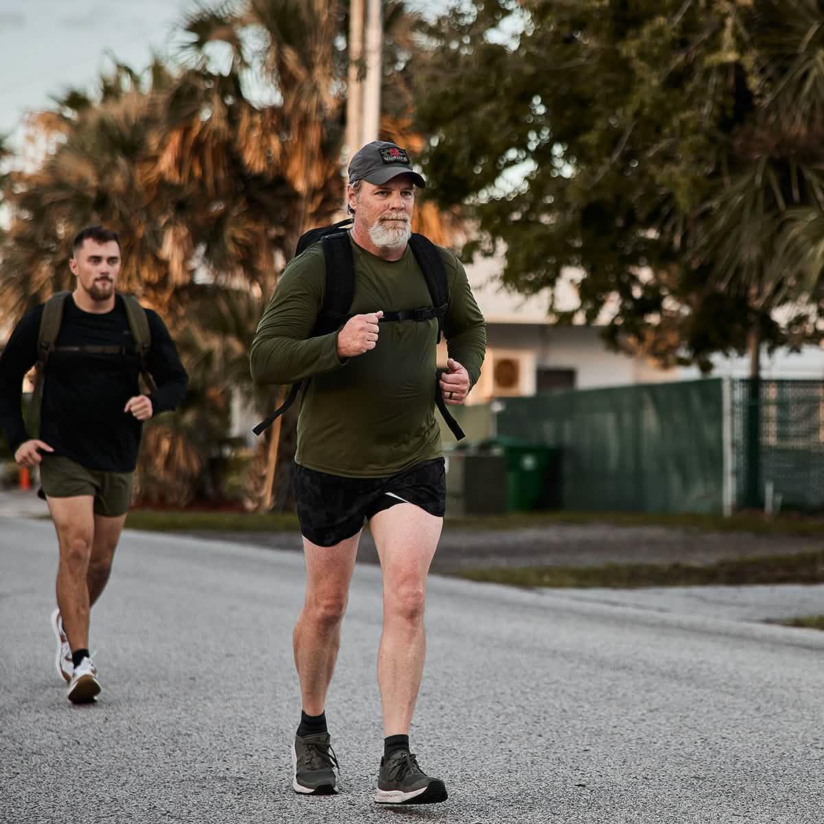 Two men rucking on a suburban street at sunset wearing backpacks and athletic clothes, surrounded by trees and fences