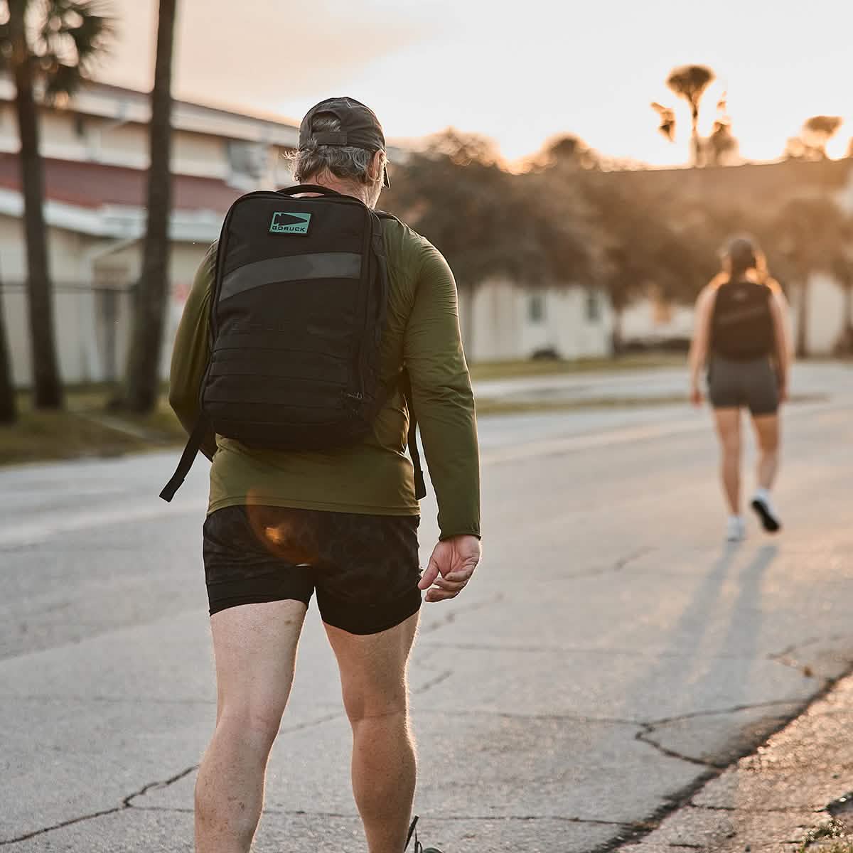 A man dressed in a green shirt and GORUCK's black Men’s Ranger Panties - ToughStretch shorts, carrying a black backpack, strolls down a sunlit street. Ahead, another individual with a backpack walks the same route. Trees and buildings outline the background beneath a clear sky.