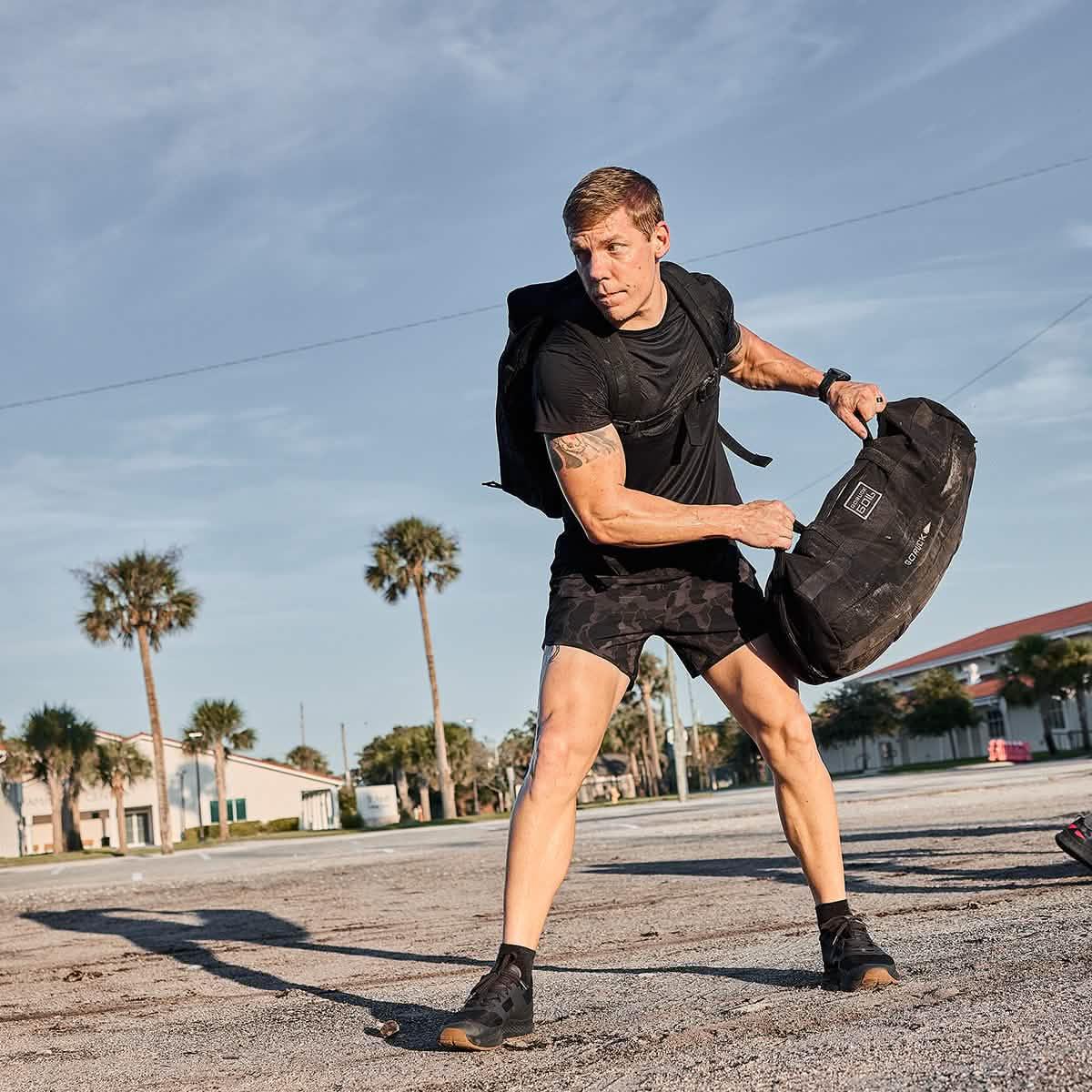 Athletic man wearing black Ranger Panties camo shorts and black shirt training outdoors with black GORUCK bag