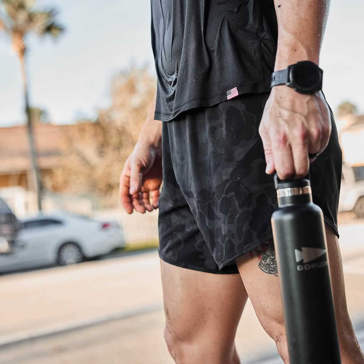 A person stands outdoors in a dark athletic outfit, showcasing the durability of GORUCK’s Men’s Ranger Panties made with ToughStretch fabric, while holding a black water bottle. A watch adorns their left wrist against a backdrop of blurred trees and cars, creating an urban street scene.