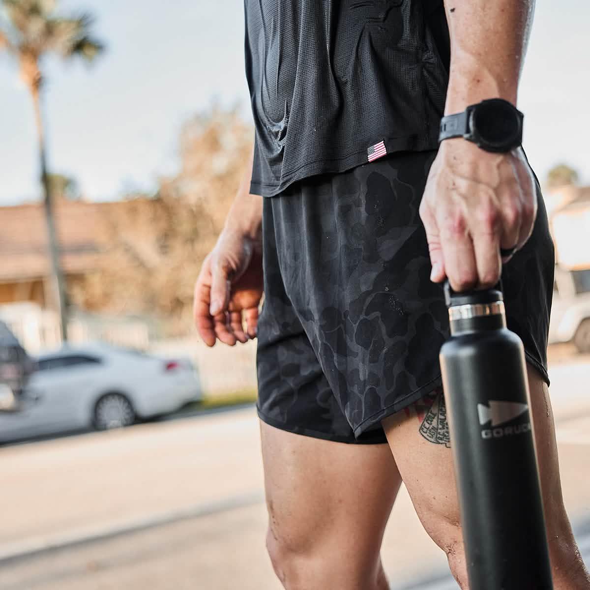 Close-up of man wearing black Frogskin camo men's Ranger shorts holding black GORUCK water bottle outdoors