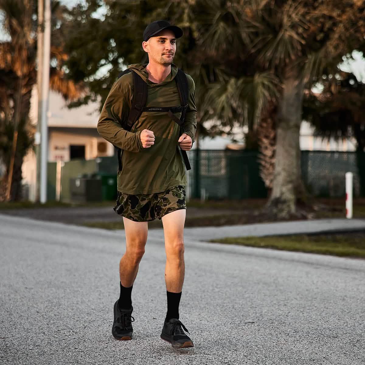 Man running outdoors wearing ranger camo shorts, olive long sleeve shirt, black cap, and backpack near palm trees
