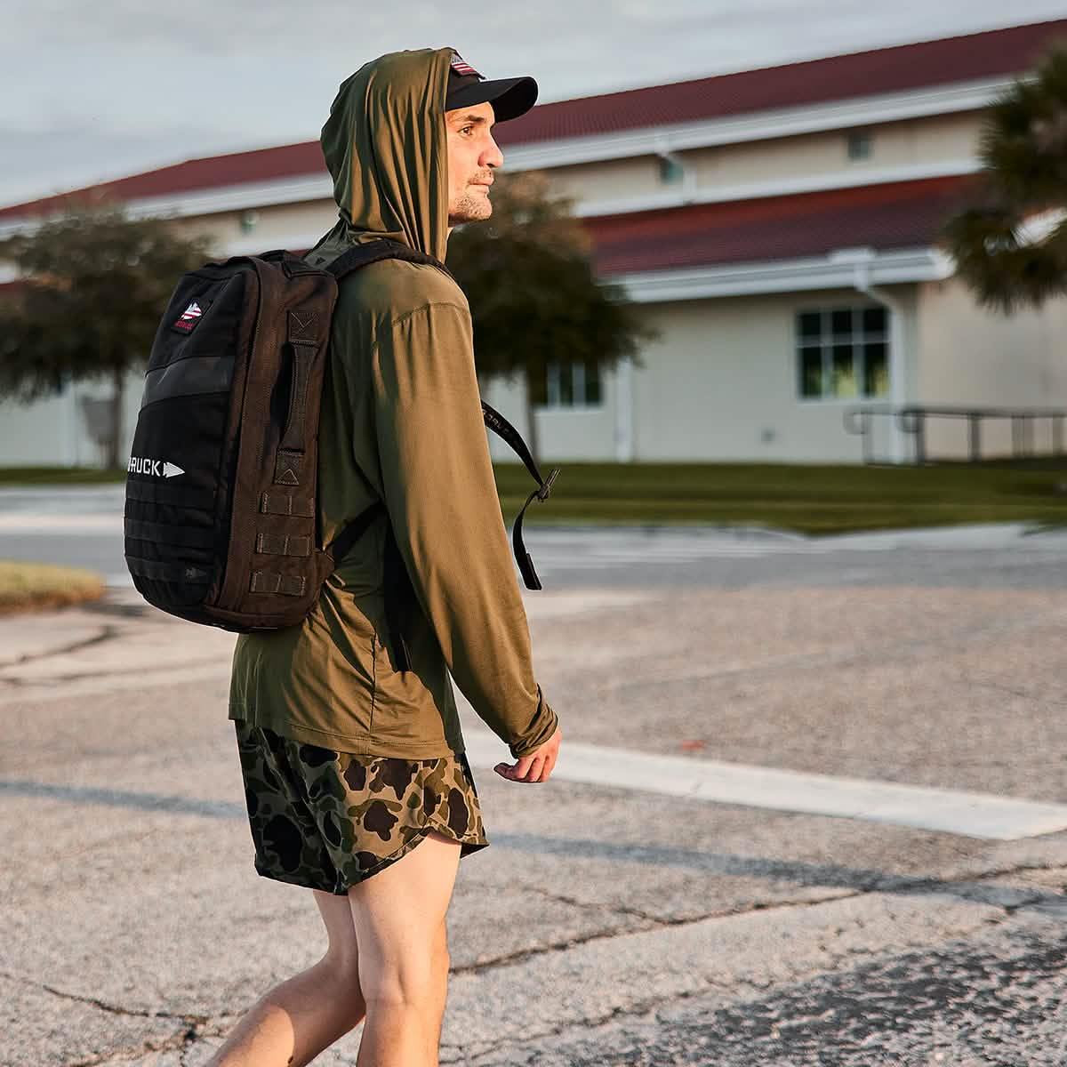 Man wearing olive hoodie and camo shorts carrying a black GORUCK backpack outdoors near building
