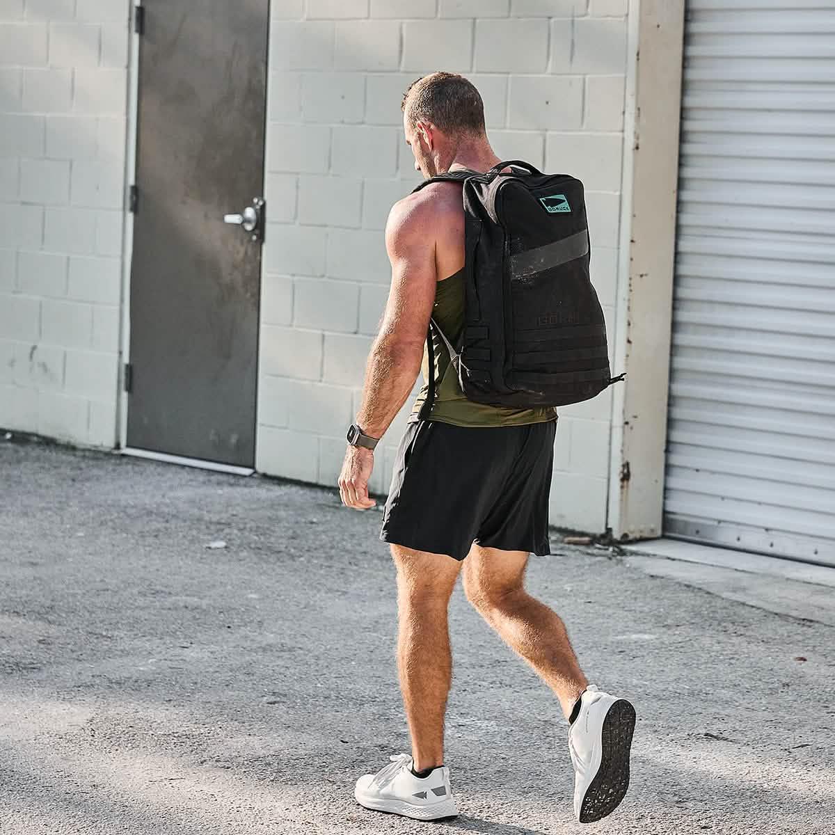 Man wearing black GORUCK backpack, black shorts, green tank top, and white sneakers walking outside an industrial building