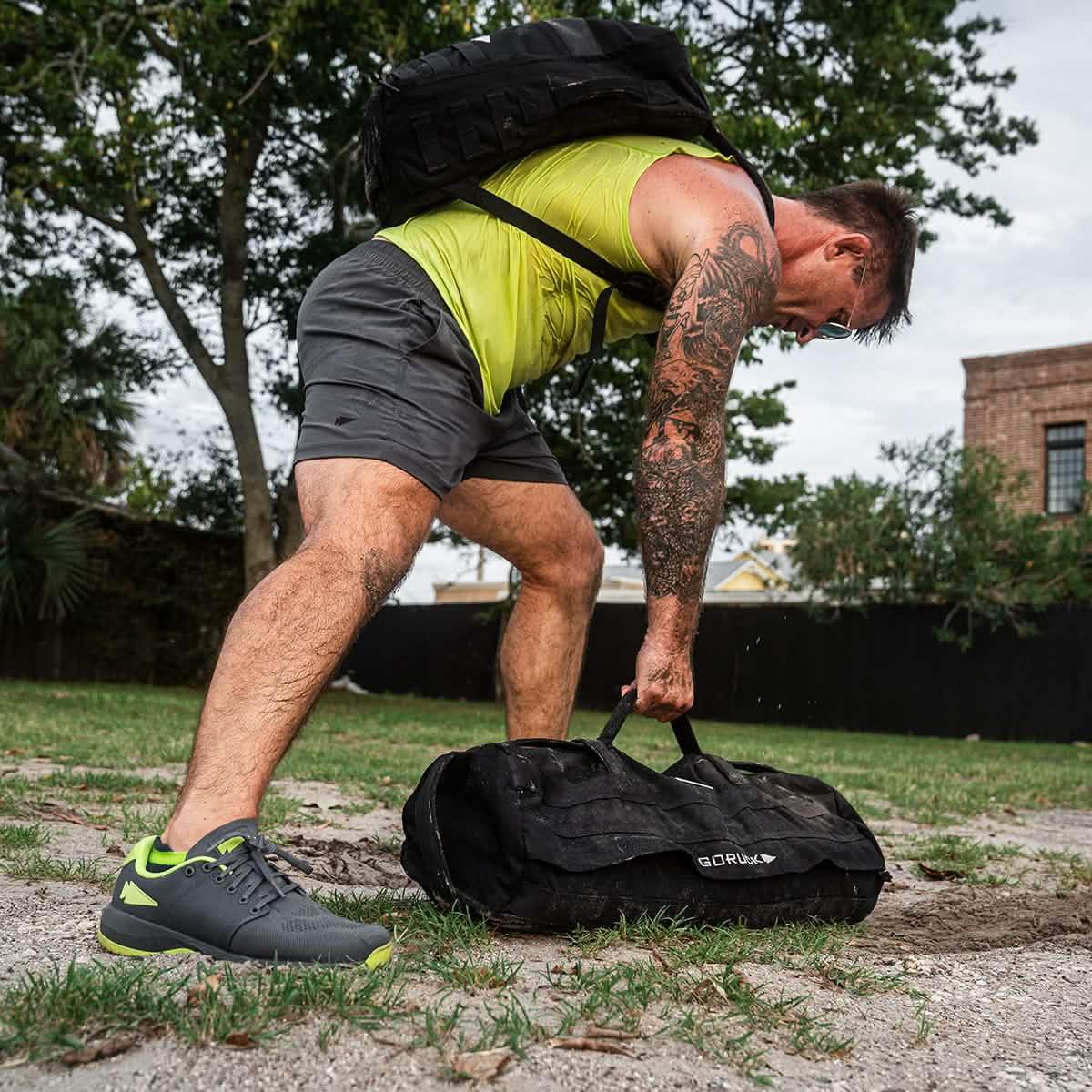 Man in neon yellow tank top and charcoal GORUCK shorts lifting a black weighted sandbag outdoors with tattooed arm