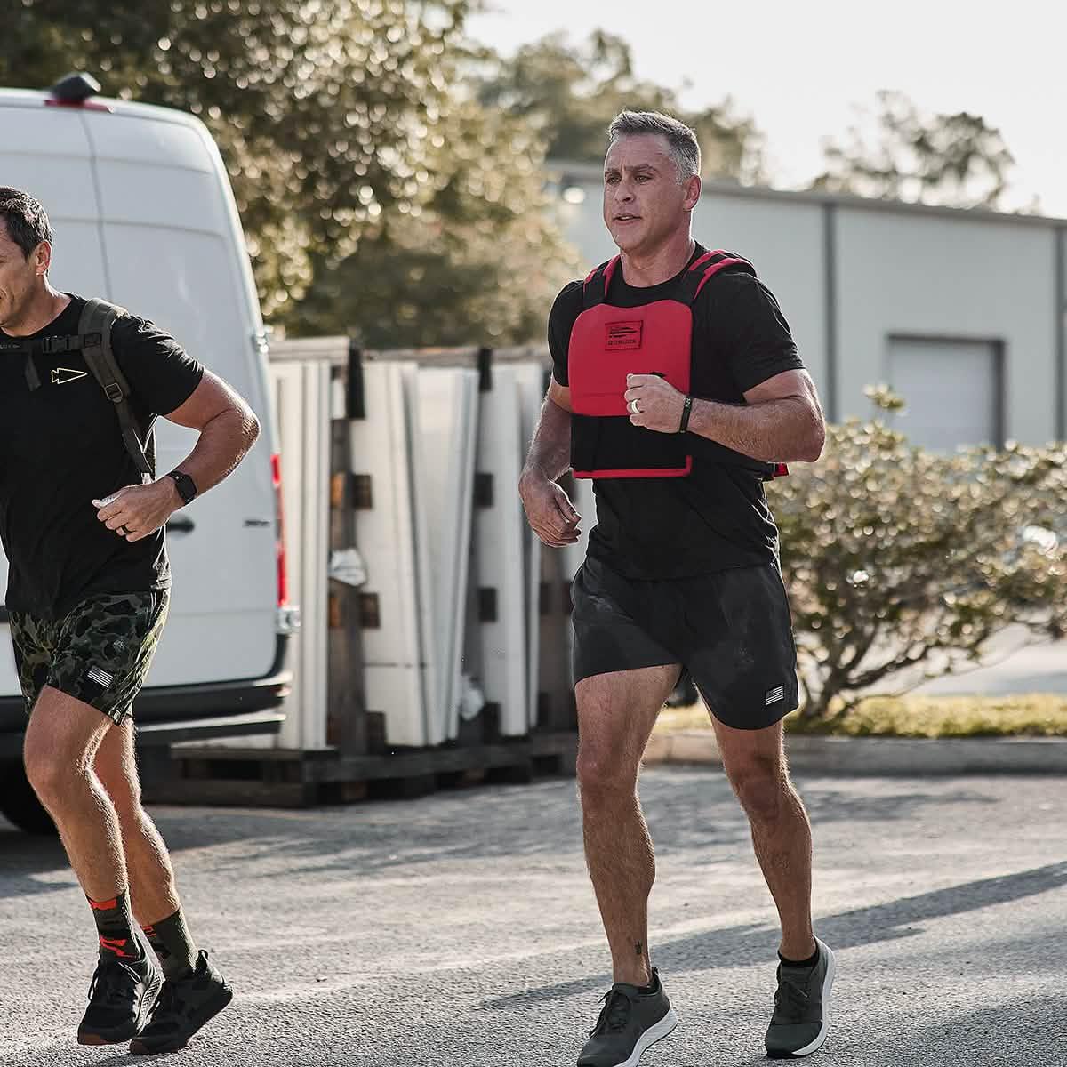 Two men running outdoors, one wearing GORUCK weighted vest and black training shorts, active fitness gear