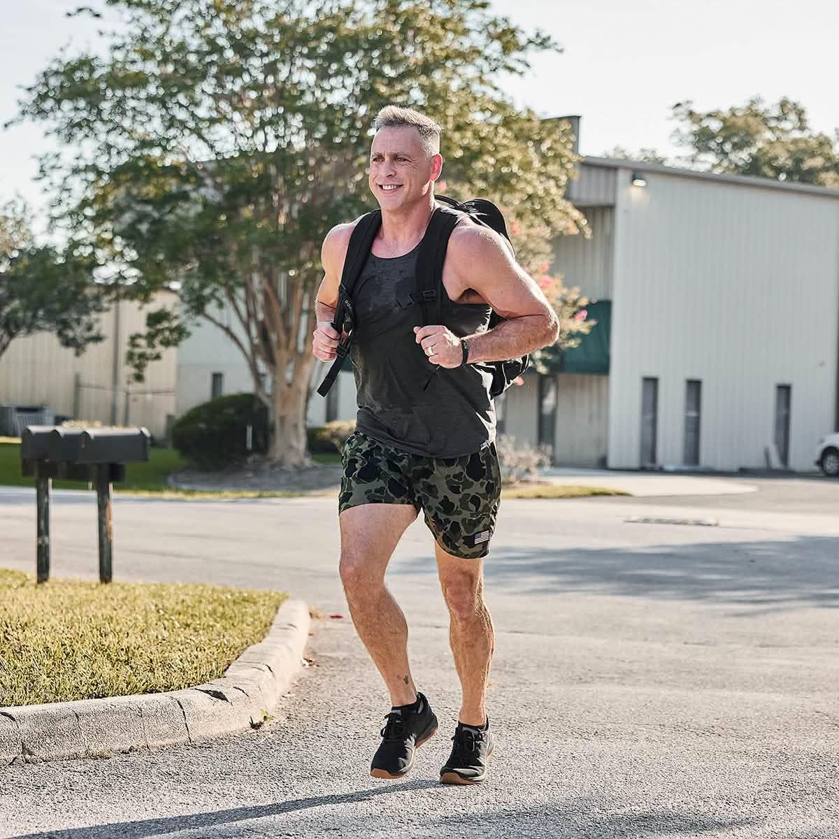 Man running outdoors wearing GORUCK camo training shorts, black tank top, and backpack in sunny urban setting