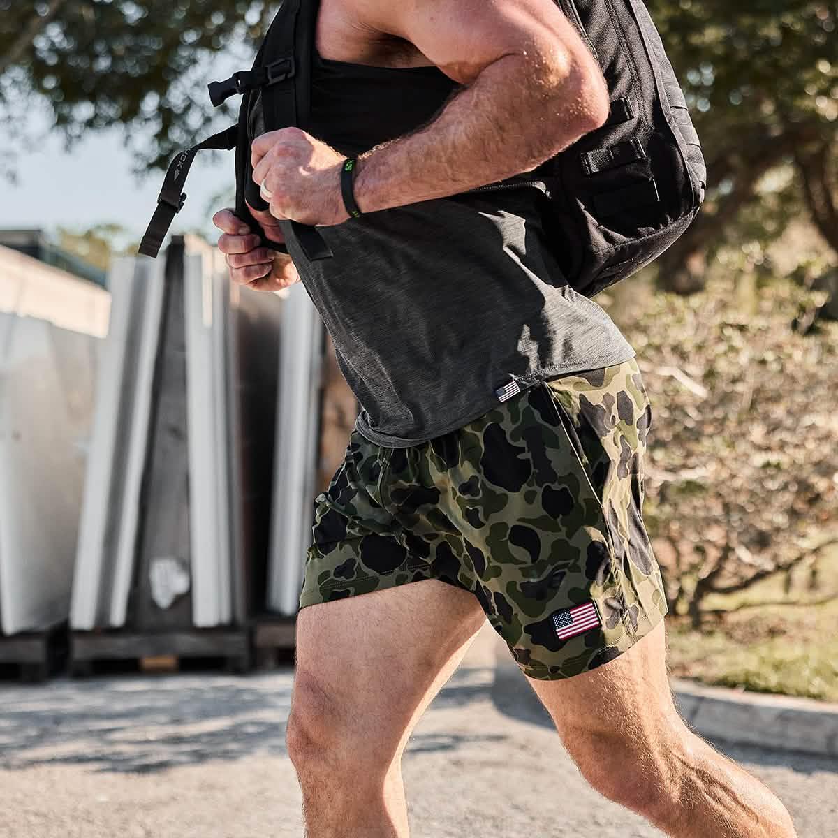 Man running outdoors wearing green ranger camo USA flag training shorts and black tank top with backpack