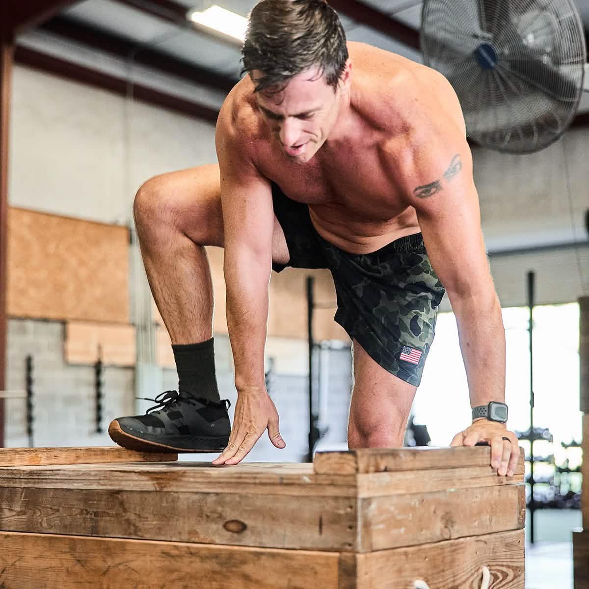 Athletic man wearing men's USA Ranger Camo training shorts with American flag patch doing plyometric box jump in gym